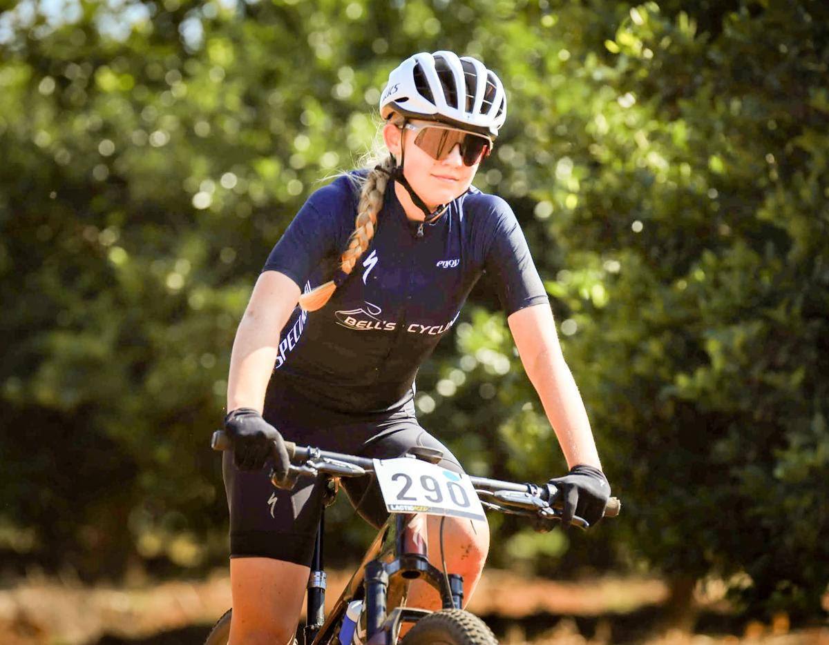A young rider in club kit and a race number, riding through dappled shade